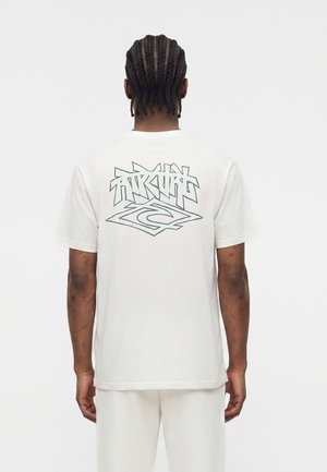 Άνδρας με πλεγμένα μαλλιά φοράει λευκό T-shirt και παντελόνι, στην πλάτη εμφανίζεται μαύρο λογότυπο σε στυλ γκράφιτι στο T-shirt.