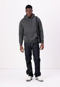 Hanorac gri cu buzunar frontal, blugi denim de culoare bleumarin și adidași albi cu dungi negre. Model purtând o geantă de umăr neagră.