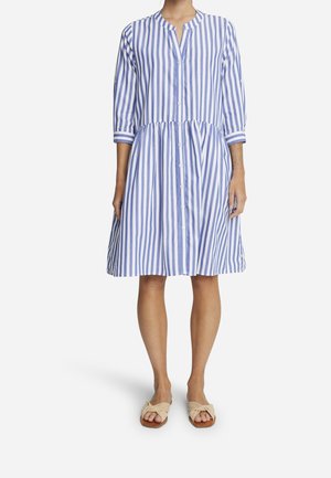 Femme portant une robe rayée bleu et blanc à rayures verticales, longueur genou, manches trois-quarts, et des sandales plates beiges.