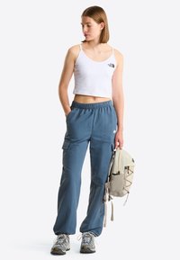Jeune femme en débardeur blanc court, pantalon cargo bleu, chaussures de randonnée, tenant un sac à dos beige, debout devant un fond uni, regardant vers la gauche.