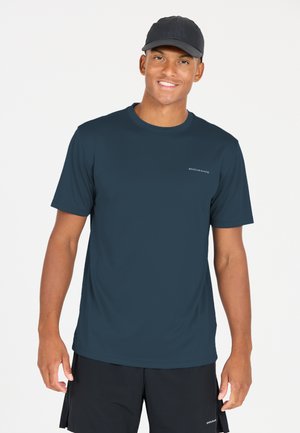 Leende man i en marinblå Endurance-t-shirt, svarta shorts och en svart keps framför en ren vit bakgrund.