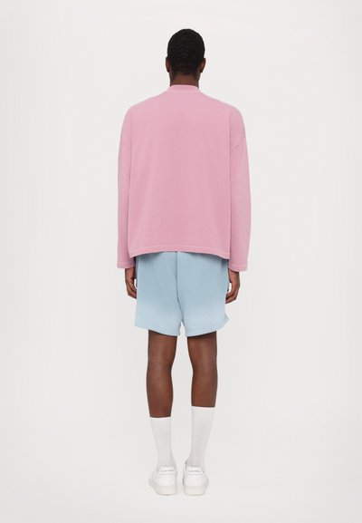 Gaiši rozā pārāk liela izmēra sporta krekls ar garām piedurknēm, kombinēts ar gaiši ziliem šortiem. Balti zeķes un sporta apavi noslēdz apģērbu.