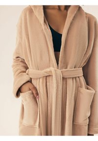 Beżowy, puszysty szlafrok z kapturem, związany w talii. Posiada dwie przednie kieszenie i miękką fakturę.