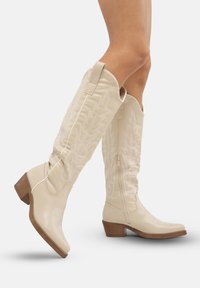 Botas western de cuero color crema hasta la rodilla, con bordados de hojas, cremallera lateral y tacón grueso. Textura suave con puntas puntiagudas.