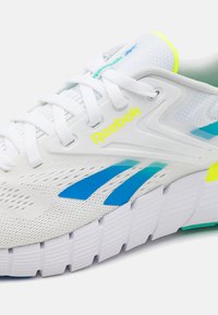 Baltas sporta apavi ar sieta materiāla augšdaļu, ziliem un zaļiem akcents, bieza balta zole ar rieviņām. Uz augšdaļas ir Reebok logo neona dzeltenā krāsā.