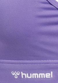 Nahaufnahme von violett geripptem Stoff mit einer Naht und weißem Hummel-Logo, das nahe am unteren Rand aufgedruckt ist.