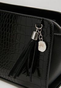 Sort faux krokodilleder taske med sølv hardware, prydet med et frynseaccent og en lille rund charme med et logo.