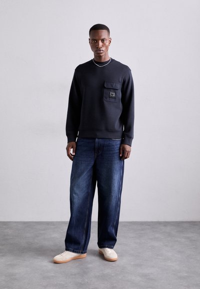 Jūrų laivyno spalvos megztinis su krūtinės kišene, derinamas su plačiomis mėlynomis džinsinėmis kelnėmis ir baltomis sportinėmis batais. Yra apvalus iškirpimas ir paramounto rankovės.