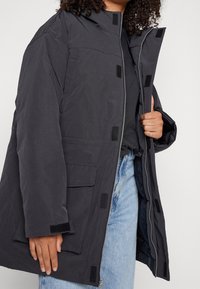 Svart vadderad jacka med hög krage, dragkedjestängning, kardborreaccents och framfickor, bärs över blå jeans. Strukturerad tyg och avslappnad passform.