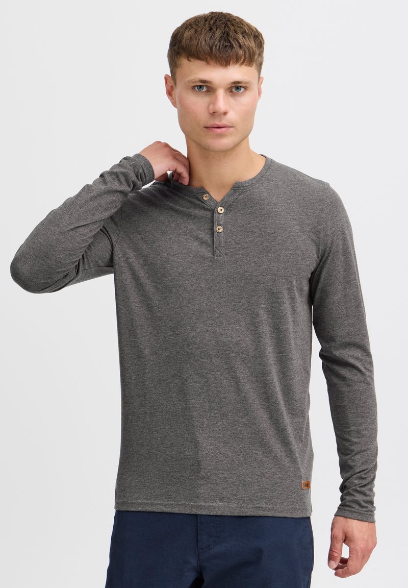 Cămașă henley lungă gri, cu mânecă lungă, fabricată dintr-un material moale, având un decolteu rotund, trei butoane din lemn și un mic patch cu logo pe tiv.
