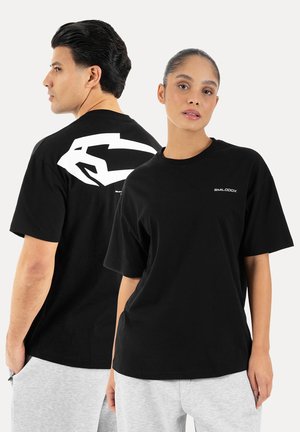 Mężczyzna i kobieta ubrani w czarne oversize T-shirty i jasnoszare spodnie, kobieta zwrócona przodem, mężczyzna obrócony bokiem, pokazujący biały wzór na plecach.