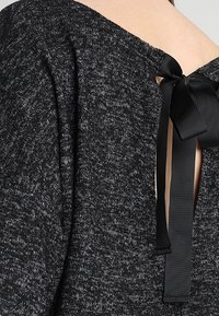 Tmavě šedý pletený svetr s širokým výstřihem a otevřením na zádech, které je upevněno černou saténovou mašlí. Měkká textura s melírovaným dezénem.