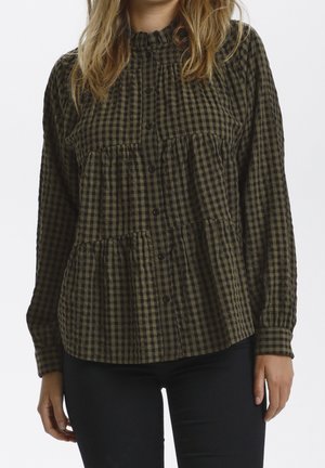 Kobieta nosząca długą, zapinaną na guziki koszulę w ciemnozieloną i czarną kratę z marszczonymi partiami i falbankowym kołnierzem, w połączeniu z czarnymi spodniami.