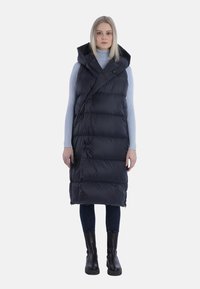 Schwarze Puffweste mit Kapuze, gestepptem Design und Wickelschluss. Getragen über einem hellblauen Rollkragenpullover, kombiniert mit dunklen Jeans und schwarzen Stiefeln.