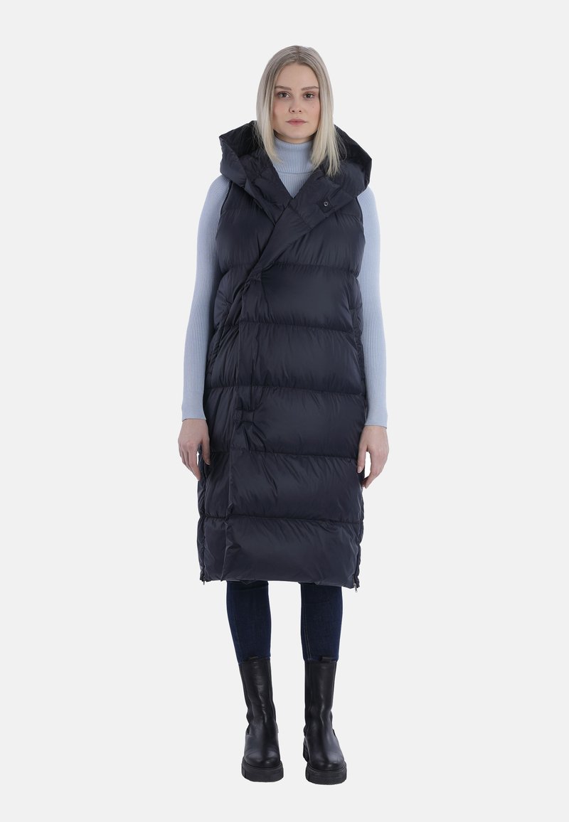 Schwarze Puffweste mit Kapuze, gestepptem Design und Wickelschluss. Getragen über einem hellblauen Rollkragenpullover, kombiniert mit dunklen Jeans und schwarzen Stiefeln.