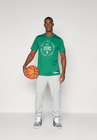 Camiseta verde de manga corta con gráfico de "Celtics Basketball" y joggers de cargo grises. El modelo sostiene un balón de baloncesto y lleva zapatillas deportivas.