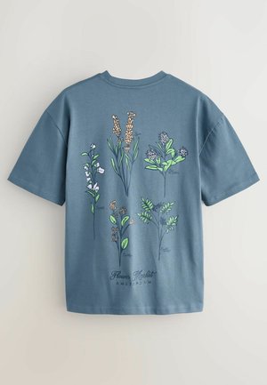 Camiseta de algodón azul con ilustraciones botánicas en verde, blanco y naranja. El texto dice "Mercado de Flores AMSTERDAM" cerca del dobladillo.