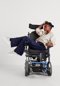 Ηλεκτρικό αναπηρικό αμαξίδιο με μαύρη καρέκλα, μπλε σκελετό και επενδεδυμένα μπράτσα. Ο χρήστης φοράει ένα ριγέ πουκάμισο και ναυτικές παντελόνες, ξεκουράζοντας το ένα του πόδι.