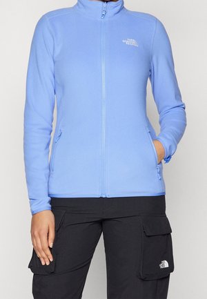 Person iført en lyseblå fleecejakke med lynlås og sorte cargobukser med The North Face-logoer på brystet og lommen.