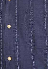 Camicia blu navy con bottoni realizzata in tessuto testurizzato, caratterizzata da righe verticali bianche, una tasca sul petto e bottoni di colore chiaro.