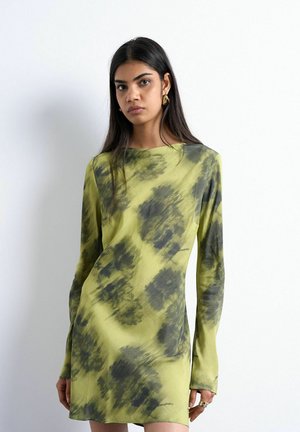 Kvinde med langt mørkt hår iført gulgrøn langærmet kjole med grå abstrakte mønstre og gyldne hængende øreringe mod en hvid baggrund.