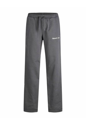 Person trägt einen grauen Kapuzenpullover und passende Jogginghose mit weißen Sneakers, steht vor einem schlichten hellen Hintergrund.