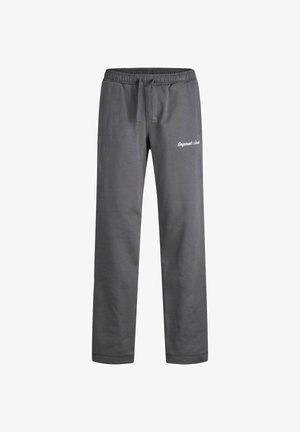 Person trägt einen grauen Kapuzenpullover und passende Jogginghose mit weißen Sneakers, steht vor einem schlichten hellen Hintergrund.