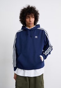 Navy blauwe hoodie met een voorzak en een witte drie-strepen ontwerp op de mouwen. Bevat een klein wit logo op de borst. Zacht materiaal.