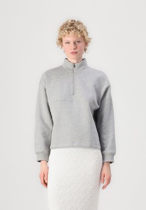 Grå hettegenser med glidelås og høy krage, avslappet passform, og ribbestrikkede mansjetter. Myk tekstur og enkelt design. Brukt over et strukturert hvitt skjørt.