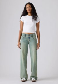 Γυναίκα με μακριά σγουρά μαλλιά που φοράει λευκό κοντό t-shirt, ανοιχτό μπλε τζιν με ευρύ παντελόνι και λευκά αθλητικά παπούτσια, μπροστά σε απλό φόντο.