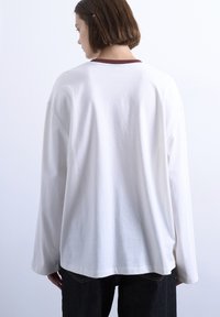 Άτομο με κοντά καστανά μαλλιά, φορώντας ένα φαρδύ λευκό πουκάμισο με μακρύ μανίκι και σκούρο παντελόνι, γυρισμένο μ ε την πλάτη σε ένα απλό ανοιχτό χρωματιστό φόντο.