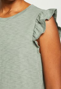 Haut vert clair à manches courtes en tissu doux, avec des manches courtes à volants et un col rond. Texture lisse avec un léger éclat.