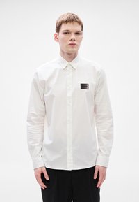 Λευκό πουκάμισο με μακριά μανίκια από απαλό βαμβάκι, με μαύρο ορθογώνιο λογότυπο στο αριστερό μέρος του στήθους, κλασικό γιακά και κούμπωμα με κουμπιά.
