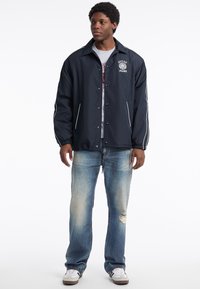 Marineblaue Windjacke mit Knöpfen, weiß abgesetzten Streifen und Logo; kombiniert mit hellblauen, verwaschenen Jeans und weißen Sneakers.