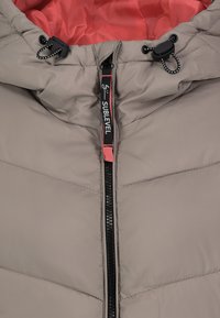 Beige pufferjack met zwarte rits, "SUBLEVEL" label, rode voering en verstelbare zwarte koorden op de capuchon.
