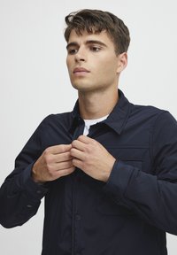 Navy blue button-up jacket with a collar, textured fabric, and a chest pocket. Model adjusts collar, revealing a white inner shirt.