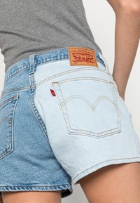 Henkilö, joka käyttää kaksivärisiä sinisiä denim Levi's 501 -shortseja harmaan paidan kanssa, näkyvissä takataskut ja nahkainen bränditarra vyötäröllä.
