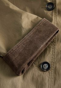 Detail blízky na khaki trenčkot, ktorý má hnedé kordúrové manžety, čierne gombíky a hladkú textúru, pričom zvýrazňuje kontrastné materiály a farby.