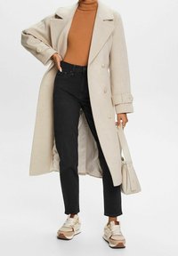 Beige wollen mantel met dubbele rij knopen over een bruine coltrui, gecombineerd met zwarte spijkerbroek en beige en bruine sneakers, geaccentueerd met een lichte tas.