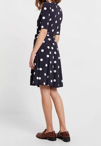 Mörkblå klänning med prickar, kortärmad och med bälten i midjan. Har en utställd kjol och vita cirkulära mönster. Bärs med bruna loafers.