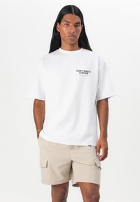 Weißes Baumwoll-T-Shirt mit kurzen Ärmeln und Rundhalsausschnitt, mit einem schwarzen Aufdruck "DON'T WASTE CULTURE" auf der Brust. Beigefarbene Cargo-Shorts.
