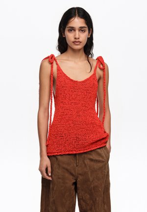 Ženska, oblečena v rdeč pleten brezrokavnik z vrvicami na ramenih in široke rjave hlače, stoji pred enobarvnim belim ozadjem.