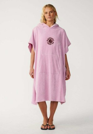 Donna che indossa un ampio vestito in spugna rosa con cappuccio, tasca frontale e infradito nere, in piedi su uno sfondo bianco semplice.