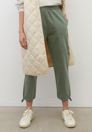 Dolna część ciała osoby noszącej oliwkowozielone, skrócone spodnie z przednią kieszenią oraz kremową pikowaną długą kamizelkę, zestawione z kremowobiałymi sportowymi butami.