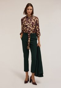Blusa dorata con motivo floreale viola, colletto annodato; abbinata a pantaloni scuri verdi su misura e scarpe con tacco alto bordeaux.