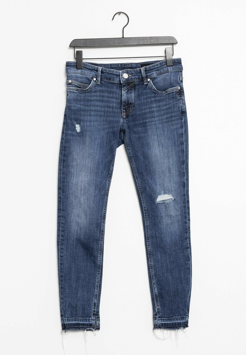 Jeans skinny en denim bleu avec une taille mi-haute, présentant un motif déchiré, des accents en métal argenté et des ourlets effilochés. Accrochés à un cintre noir.