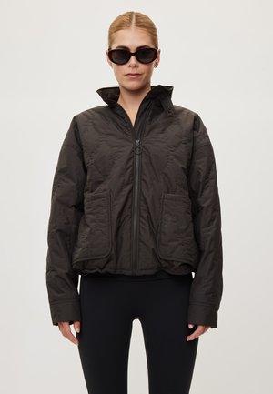 Femme portant une veste matelassée noire avec poches avant et col zippé, des leggings noirs et de grandes lunettes de soleil noires, debout sur fond blanc.