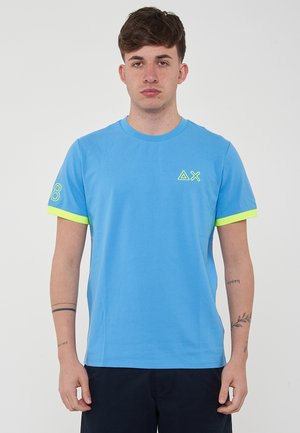 T-shirt azzurro chiaro in cotone, con maniche corte decorate da accenti giallo neon e un logo grafico sul petto e sulla manica.