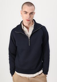 Sudadera con cremallera en color navy, con cuello y dobladillo acanalados, que presenta un logo sutil. Lleva puesta sobre una camisa de cuello a rayas beige y blanca.