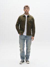 Olivovo zelená bunda s dvoma náprsnými vreckami, nosená cez bielu košeľu. Svetlomodré rifle s miernym efektom vyprania, kombinované s čierno-bielymi teniskami.